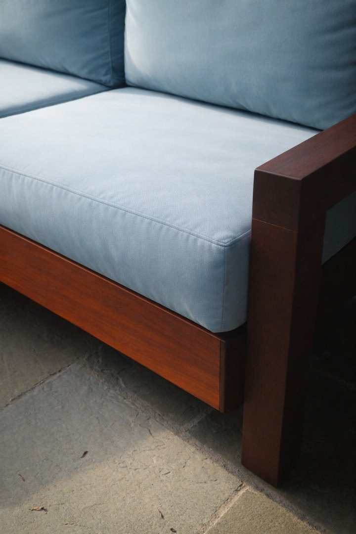 Close-up of a light blue outdoor cushion on a mahogany-toned wood frame, on a stone patio