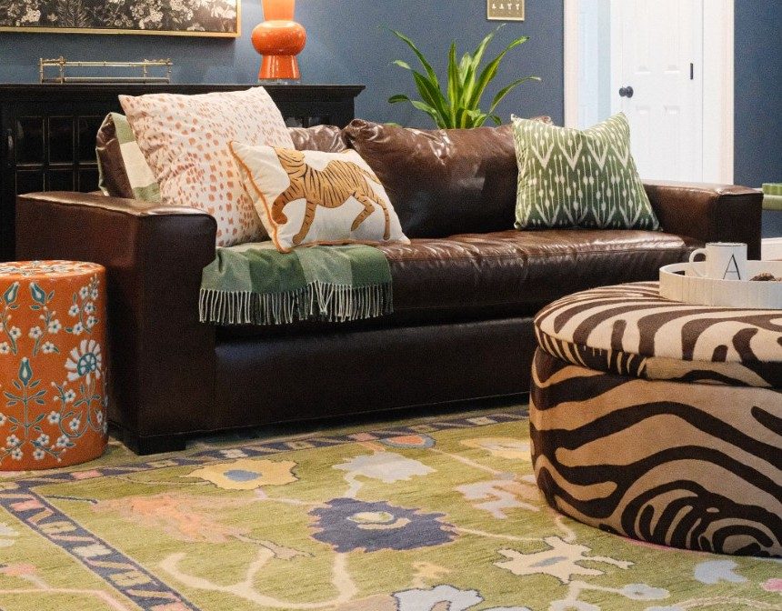 Dark brown leather sofa with patterned pillows and a green fringed throw beside a zebra-print ottoman