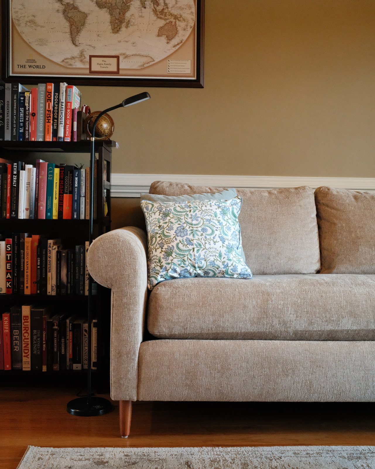 Oatmeal chenille sofa with a floral pillow beside a bookshelf and floor lamp