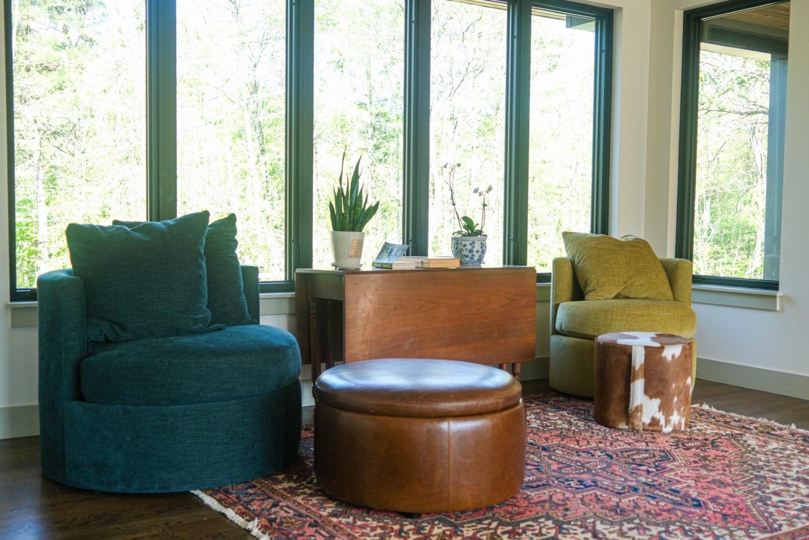 Teal and olive round lounge chairs with a leather ottoman and a cowhide pouf before tall green-framed windows