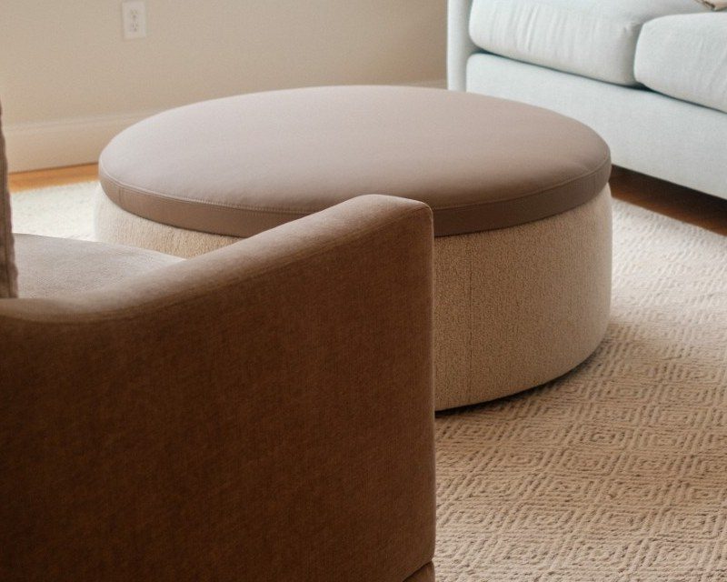 Large round ottoman with a taupe leather top and oatmeal fabric base beside a pale blue sectional